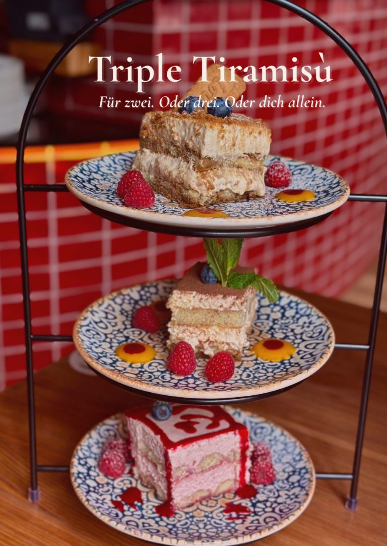 Triple Tiramisu auf einer dreistöckigen Etagere mit drei verschiedenen Tiramisu-Variationen, angerichtet auf blau gemusterten Tellern mit frischen Himbeeren und Beerensauce im Restaurant Pizzazza in München